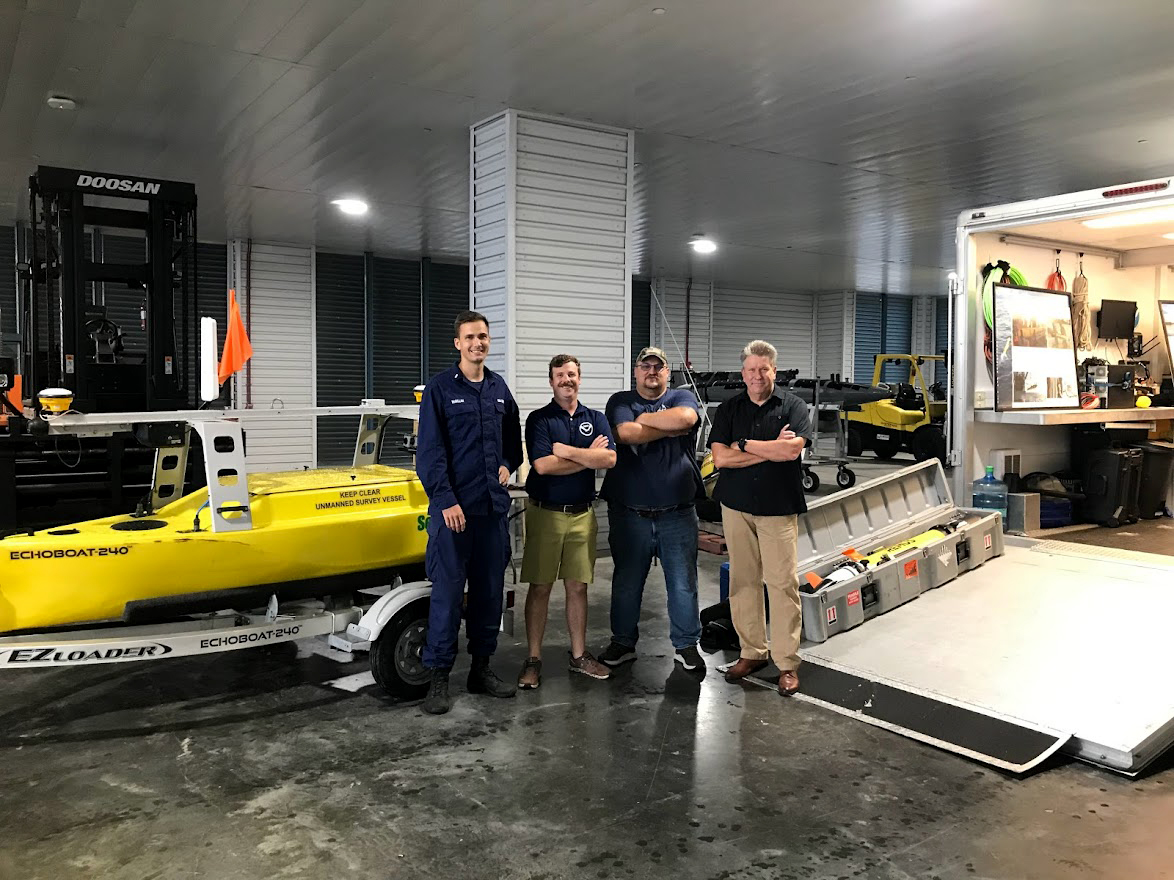 An image of (left to right) LTJG Collin McMillan, Alex Ligon, Joshua Bergeron, and Tim Osborn standing in front of the Echoboat uncrewed surface vessel.