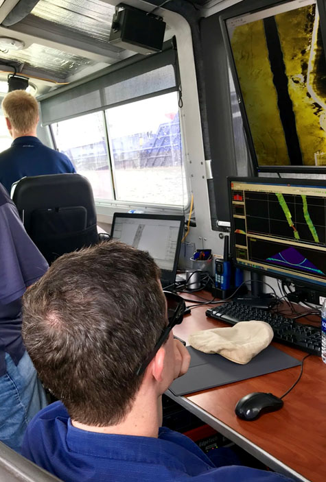 Michael Bloom drives while Lt. j.g. Patrick Lawler monitors survey acquisition. Joshua Bergeron reviews side scan data collected from the previous day looking for potential dangers to navigation.