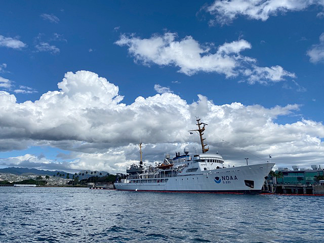 This image shows the survey vessel NOAA Ship Rainier docked alongside Ford Island which is located in Pearl Harbor, Hawaii. The image was taken on a mostly clear day in February 2022 with several white clouds in the sky.