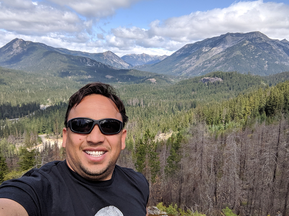 An image of Fernando Ortiz shown with a background of evergreen forests and mountains on a partly cloudy day.