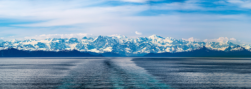 An image of the wake of a cruise ship in Prince William Sound with snow-covered mountains in the background.