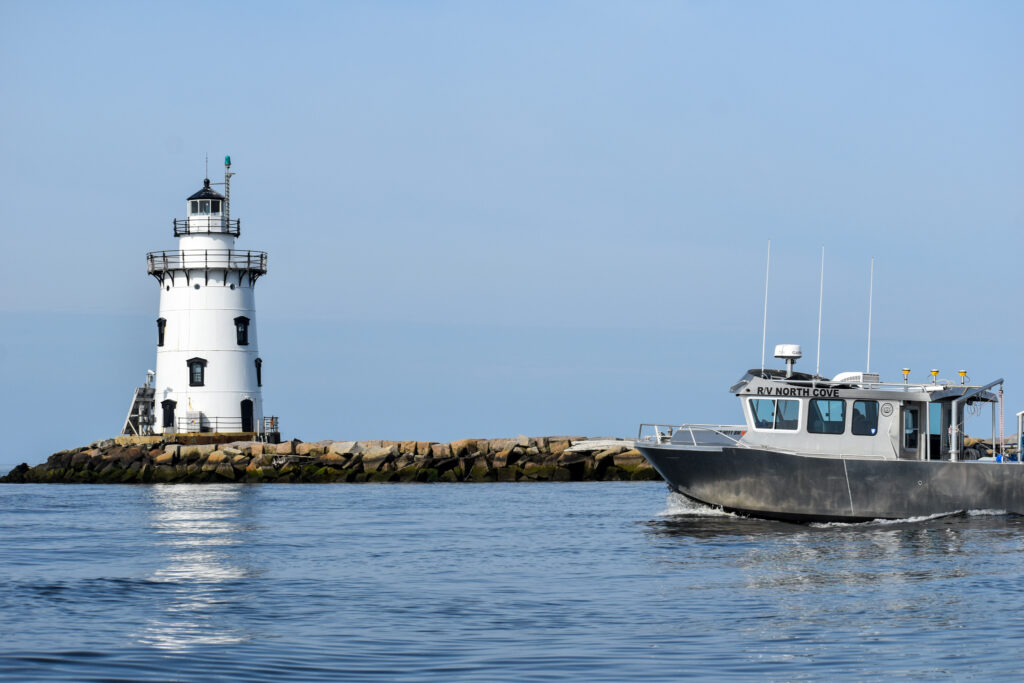 Image of a white lighthouse on a rocky shore. To the right, a silver research vessel collects hydrographic survey data in the water.